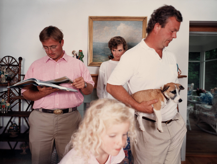 A candid family scene in a living room with three adults and a child. On the left, a man in a pink shirt is engrossed in reading a magazine. In the center, a woman peeks over the shoulder of a man on the right, who is holding a small brown and white dog. In the foreground, a young girl with blonde hair is partially visible, her face turned away from the camera. The room is decorated with a painting, a spinning wheel, and various knick-knacks, suggesting a cozy, lived-in space.