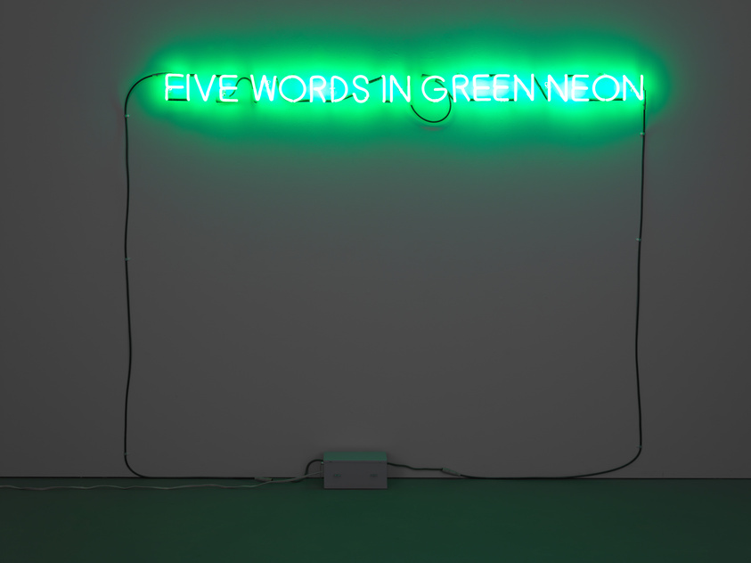 A green neon sign mounted on a grey wall with the text "FIVE WORDS IN GREEN NEON" illuminated in bright green, with visible wires and a transformer box connected to the sign at the bottom.