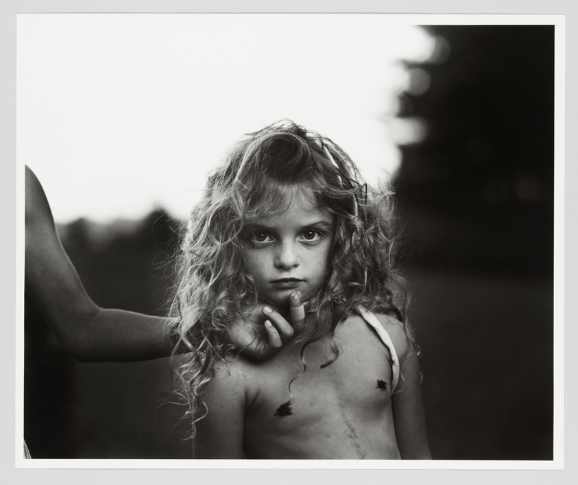 A black and white photograph of a young girl with curly hair looking directly at the camera with a solemn expression. She has leaves stuck to her chest and arm, and an adult's hand is visible on the left side of the frame, gently touching her shoulder. The background is softly blurred with natural tones.