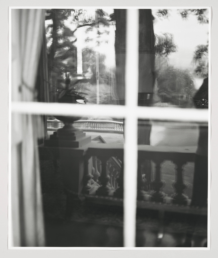 Black and white photograph looking through a window pane with reflections, showing a blurred view of trees and a balustrade outside, with a glimpse of an interior room's edge and curtain to the left.