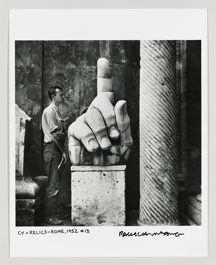 A black and white photograph featuring a large, fragmented marble hand with an extended index finger, part of an ancient statue, next to a standing man in profile who appears to be observing the relic. The background shows weathered walls, and the image includes inscriptions at the bottom indicating the subject and location as relics in Rome, dated 1952.