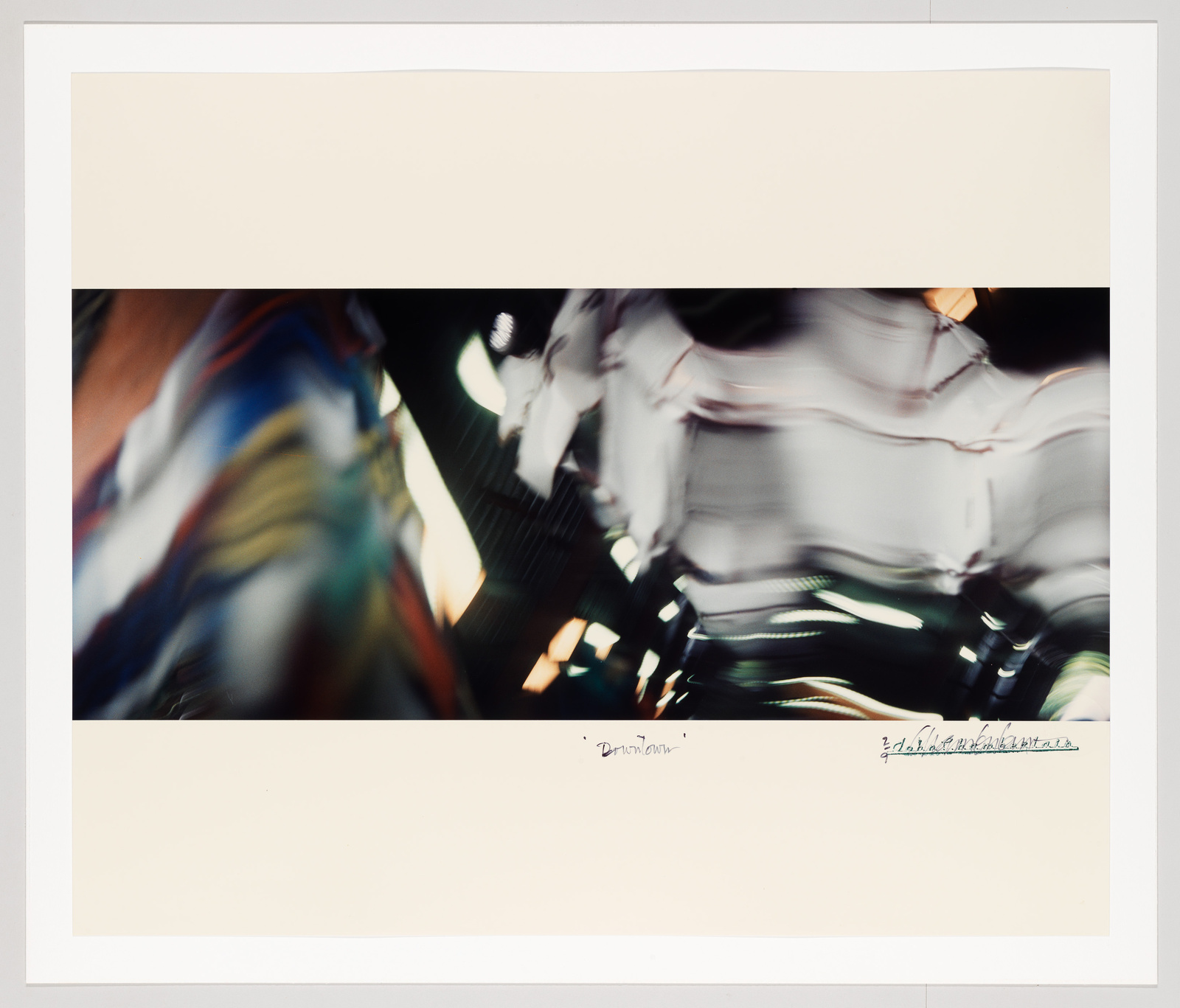 A blurred photograph capturing motion, possibly in an urban setting, with streaks of white and colored lights against a dark background, suggesting movement and speed. The image is bordered by a wide white margin with handwritten inscriptions at the bottom.