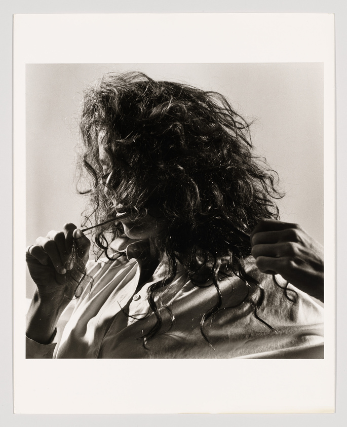A black and white photograph capturing a person with curly hair obscuring their face, holding sunglasses in one hand near their chin. The individual is wearing a light-colored shirt, and the lighting creates a dramatic contrast, highlighting the textures of their hair and shirt.