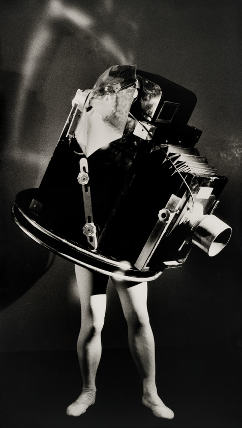 A surreal black and white photo of a person with a large camera for a head, standing against a shadowy background.