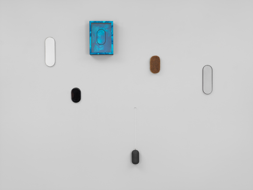 A minimalist art installation on a white wall featuring a symmetrical arrangement of seven objects: two elongated white shapes, two black oval objects, a brown textured piece, a blue framed artwork with a circular motif, and a black object hanging from a string.
