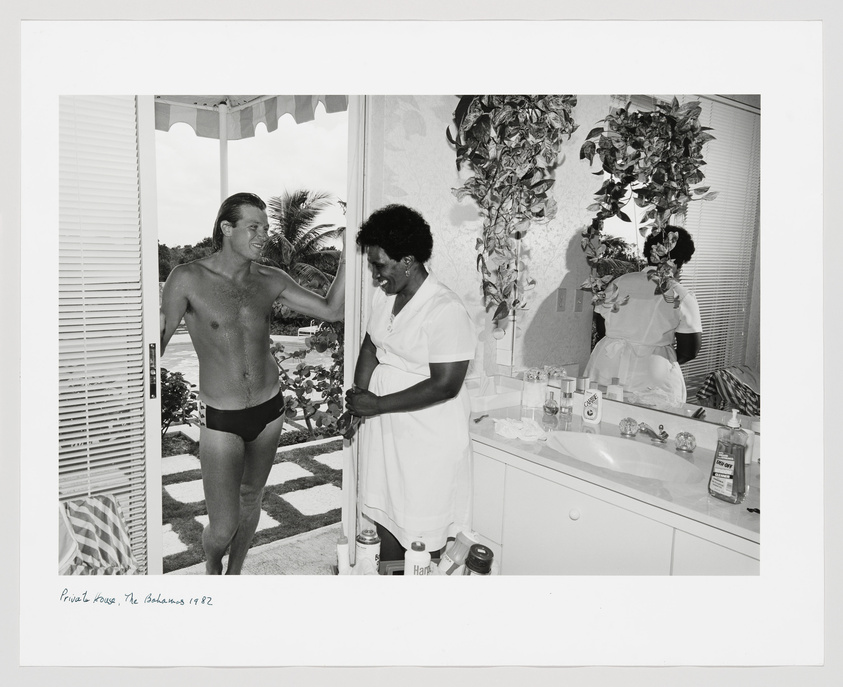 A black and white photograph depicting a shirtless man in swim trunks standing in a doorway, smiling and conversing with a woman in a white uniform, possibly a housekeeper or attendant. The setting appears to be a luxurious bathroom with a large mirror reflecting the woman's back, and various toiletries are visible on the countertop. The image has a caption at the bottom that reads "Private House, The Bahamas 1982."