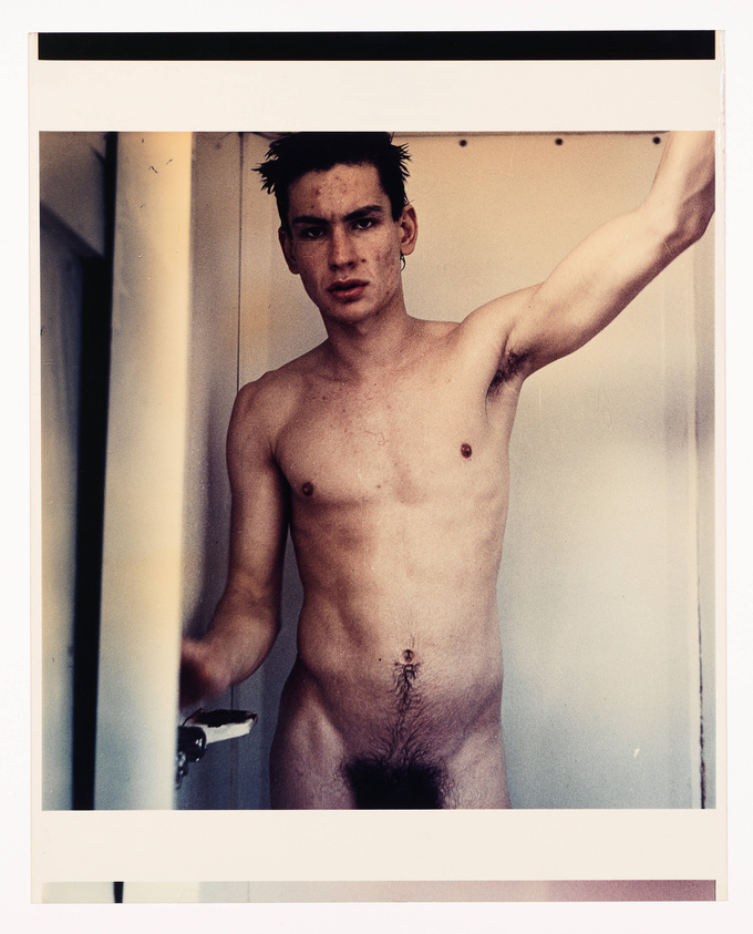 A young man with a bare chest stands in a doorway, holding the door open with his right arm extended. He has dark, spiky hair and a serious expression on his face. The photo has a vintage, polaroid-style border.