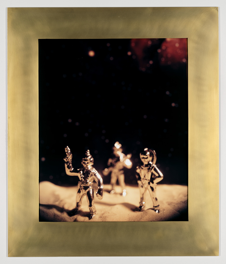 A staged photograph featuring three small, metallic astronaut figurines on a sandy surface, with a dark background speckled with white dots resembling stars. The image is framed with a yellowish border.