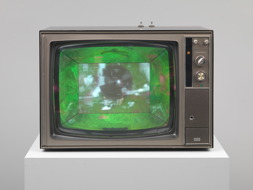 A vintage television set with a green tinted screen displaying a blurry black and white image is placed on a white pedestal against a white wall. The TV has a dark brown body with silver accents and dials on the right side.