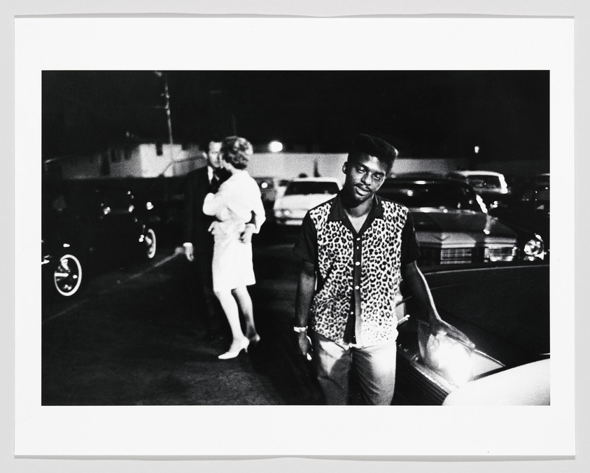 A black and white photograph capturing a moment on a street at night. In the foreground, a man in a leopard print shirt and dark pants stands beside a car, looking towards the camera with a slight smile. In the blurred background, a couple appears to be embracing near other parked cars under streetlights.