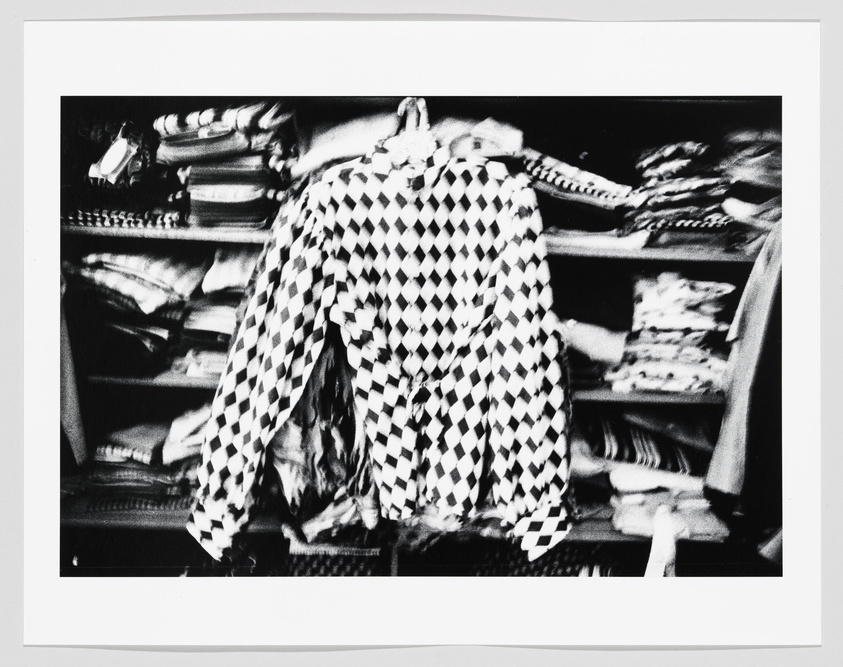 A black and white photograph capturing a person from behind, wearing a checkered jacket, in front of a blurred background featuring shelves stocked with various fabrics or clothing items. The motion blur suggests movement, either of the subject or the camera during the shot.