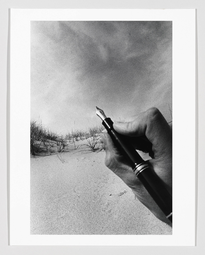 A black and white photograph featuring a person's hand holding a fountain pen, with the pen tip touching the sandy ground. Sparse vegetation is visible in the background under a cloudy sky.