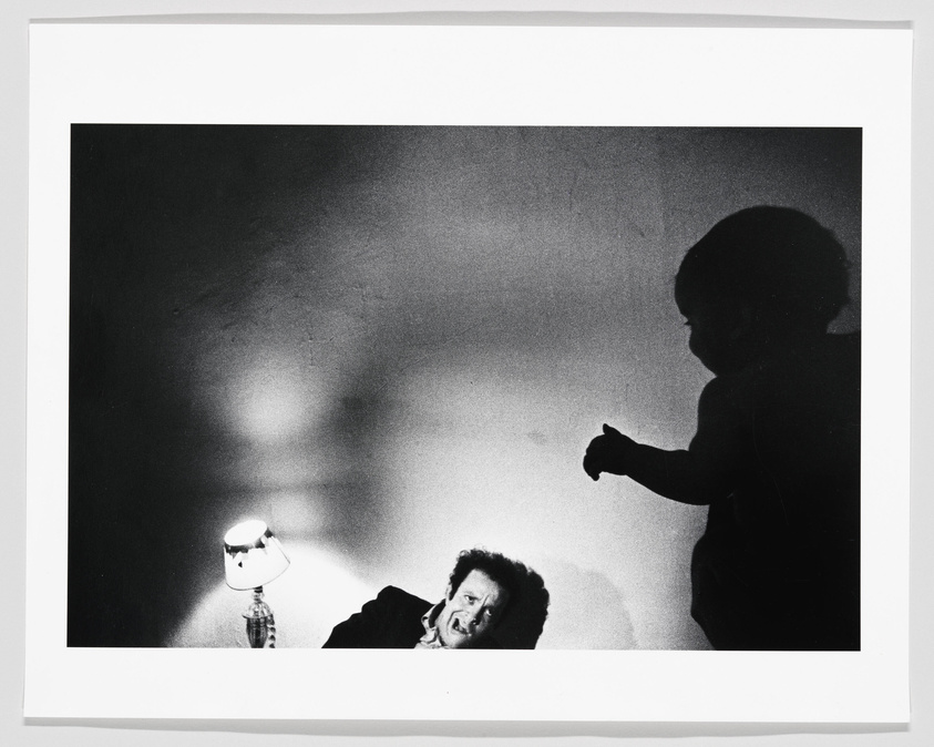 A black and white photograph capturing the silhouette of a small child pointing towards a man who is lying down, appearing to be frightened or surprised. The man's face is illuminated by a lamp with a checkered lampshade, casting dramatic shadows on the wall behind him. The image has a stark contrast between light and dark areas, creating a suspenseful atmosphere.