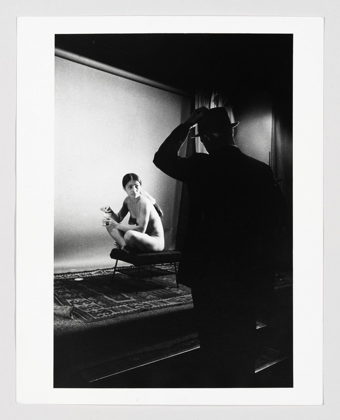A black and white photograph capturing a moment between a photographer and a model. The model is seated on a bench, looking towards the camera with a contemplative expression, while the photographer, in silhouette, is framing a shot with his hands. The setting appears to be a simple room with a plain backdrop and a patterned rug on the floor.