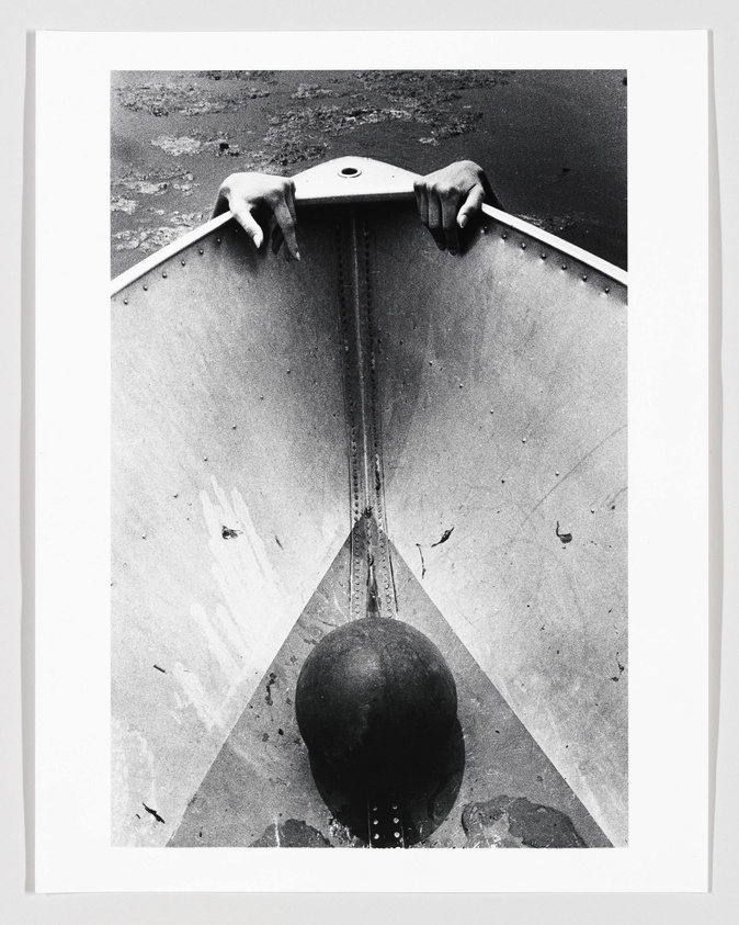 A black and white photograph showing a person's hands gripping the top edge of a metallic, V-shaped structure with a round object at the bottom, resembling a pendulum or wrecking ball, against a textured background.