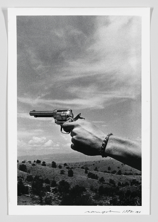A black and white photograph depicts a close-up of a person's arm extended, holding a revolver against a landscape backdrop with scattered trees and a cloudy sky. The image has a signature and date at the bottom right corner.