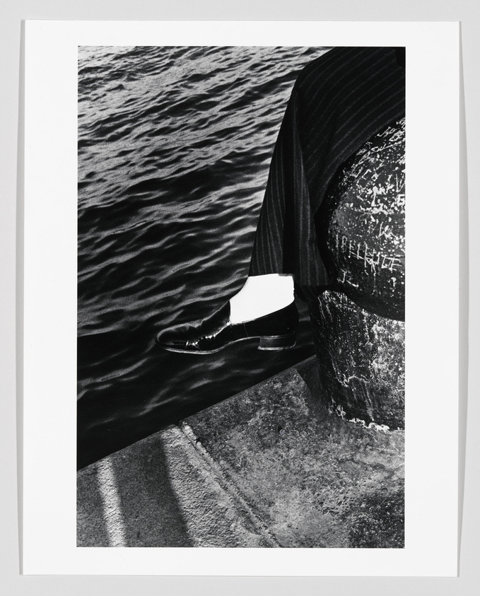 A black and white photograph capturing a close-up of a person's lower body, dressed in a pinstripe suit, standing on a concrete edge next to water. The person's polished shoe is dangling just above the rippling water surface, and the texture of the concrete is visible in the foreground. The image has a high contrast between the dark clothing, the glistening water, and the rough texture of the concrete.