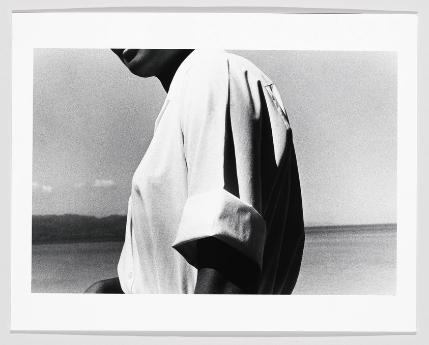 Black and white photograph of a person viewed from the side, focusing on the draped fabric of their garment with a blurred landscape in the background.