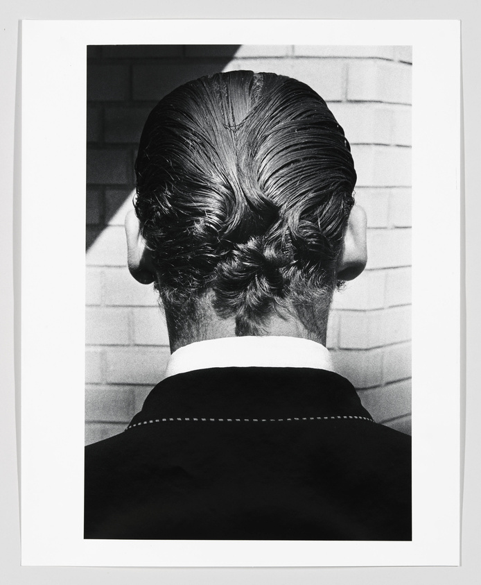 Black and white photograph of the back of a person's head, showing a neatly combed and intricately braided hairstyle. The person is wearing a dark jacket with a white collar, and is standing against a background of light-colored bricks.