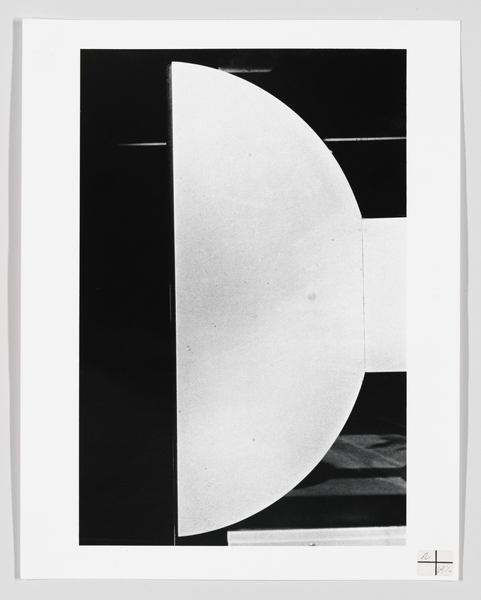 A black and white abstract photograph featuring a stark contrast between geometric shapes, with a prominent half-circle on the right side against a dark background. The image includes a signature in the lower right corner.