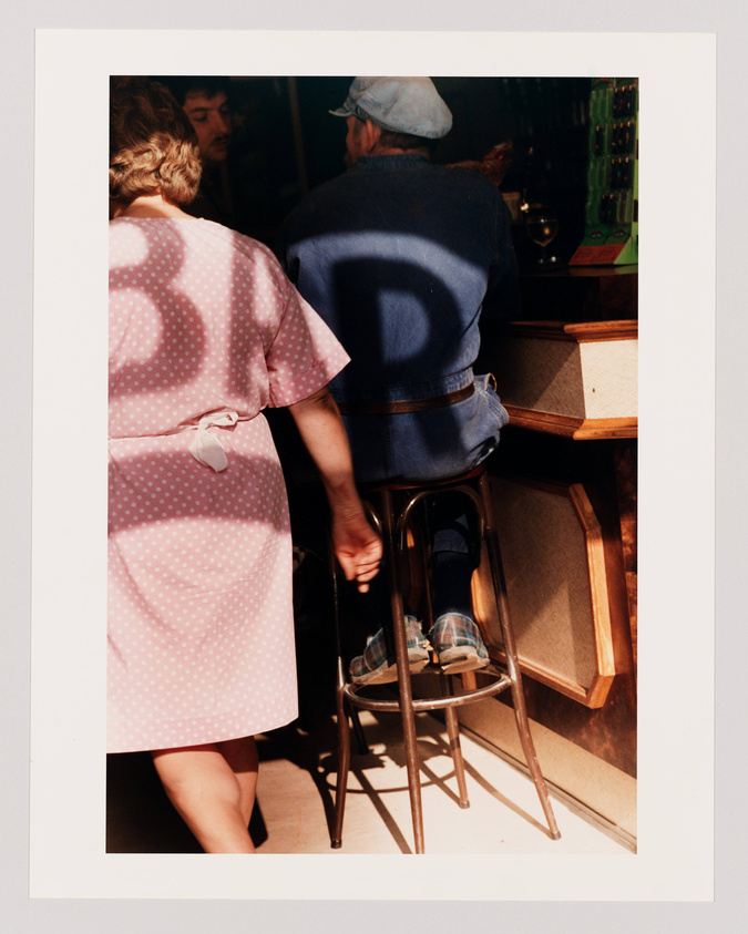 A photograph capturing a candid moment where a woman in a pink polka dot dress stands next to a man seated on a bar stool wearing a denim jacket and a cap. The man is facing a bar counter with a reflection visible in the mirror behind it, and there's a glass of white wine on the counter. The scene is bathed in warm sunlight, casting a shadow with a distinct "D" shape onto the man's back.
