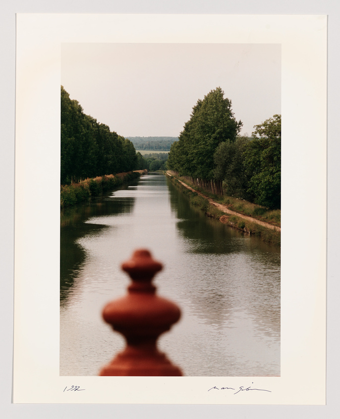 A photograph featuring a serene canal flanked by rows of trees, with the perspective taken from behind a blurred fence post in the foreground. The image is framed with a white border, signed and numbered at the bottom.