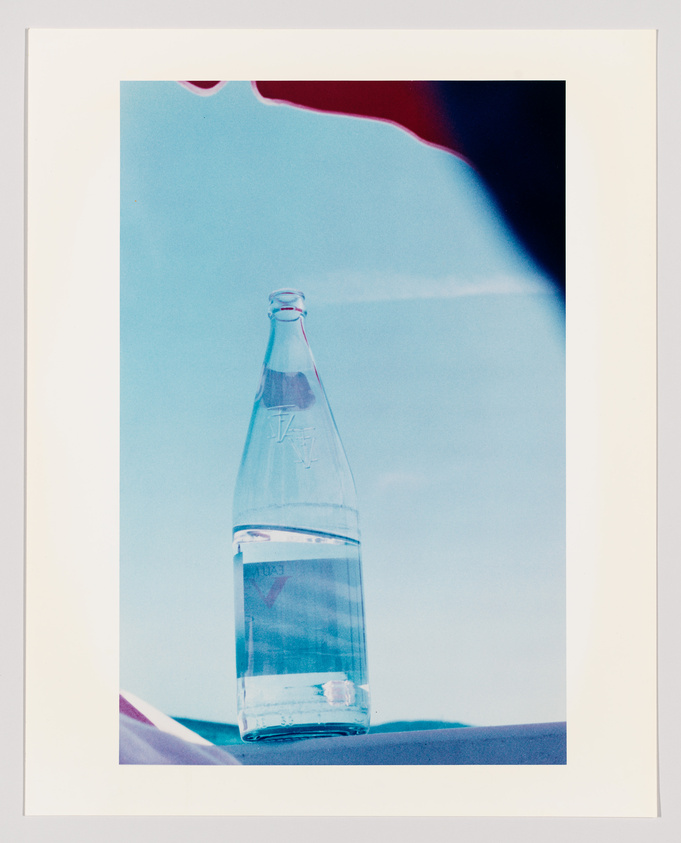 A clear glass bottle filled with water is centered against a blue background with a red and white gradient at the top corner, suggesting a reflection or light effect. The bottle appears to have text or a logo embossed on its surface.