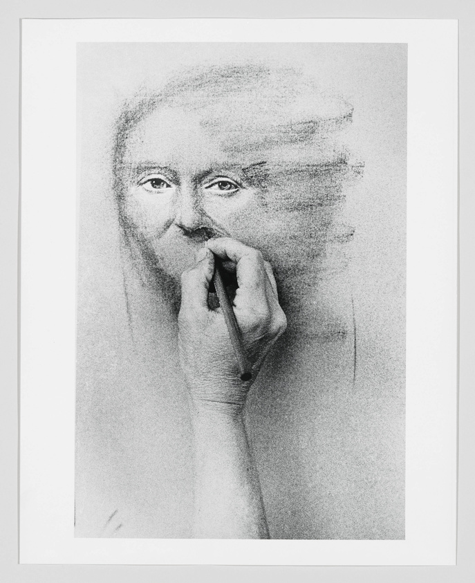 A black and white artwork depicting a hand drawing the face of a woman, with the drawing hand and pencil appearing as a continuation of the woman's portrait, creating an illusion as if the woman is drawing herself. The image has a sketch-like quality with visible pencil strokes and shading.