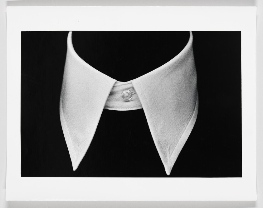 A high-contrast black and white photograph of a shirt collar and a button, framed against a black background with a white border around the image.