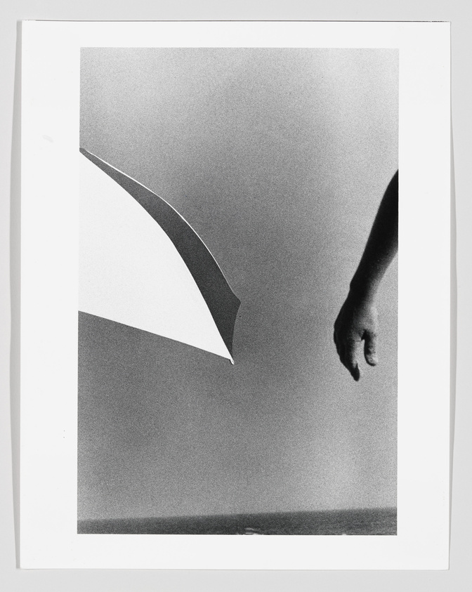 A black and white photograph featuring an abstract composition with a partial view of a human hand reaching towards a geometric shape resembling a curved sail or wing. The background is a gradient of light to dark tones, creating a sense of depth and movement.