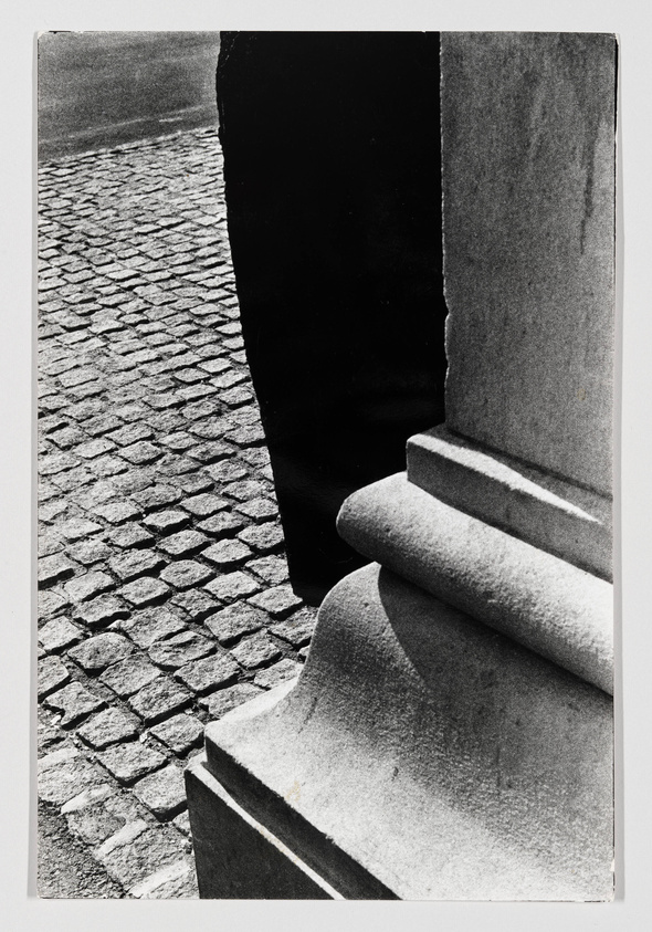 Black and white photograph capturing a close-up view of a cobblestone street with the edge of a building and part of a column base in the foreground, highlighting textures and contrast between light and shadow.