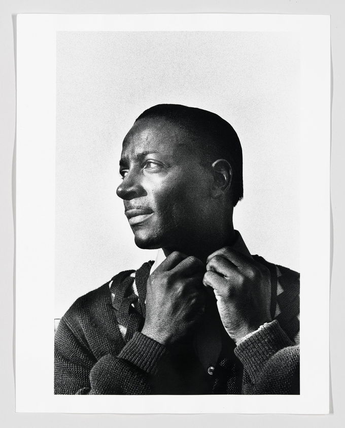 Black and white portrait of a man in profile looking to the right with a contemplative expression, adjusting the collar of his textured sweater.