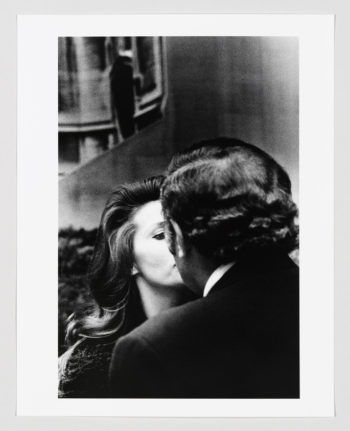 A black and white photograph capturing a close-up of a man and a woman in a romantic embrace, with the man's back to the camera and the woman's face partially obscured by his head. The focus is on the woman's visible eye and the man's hair, creating a sense of intimacy and privacy. The background is out of focus, with a hint of a reflective surface, possibly a mirror, adding depth to the image.