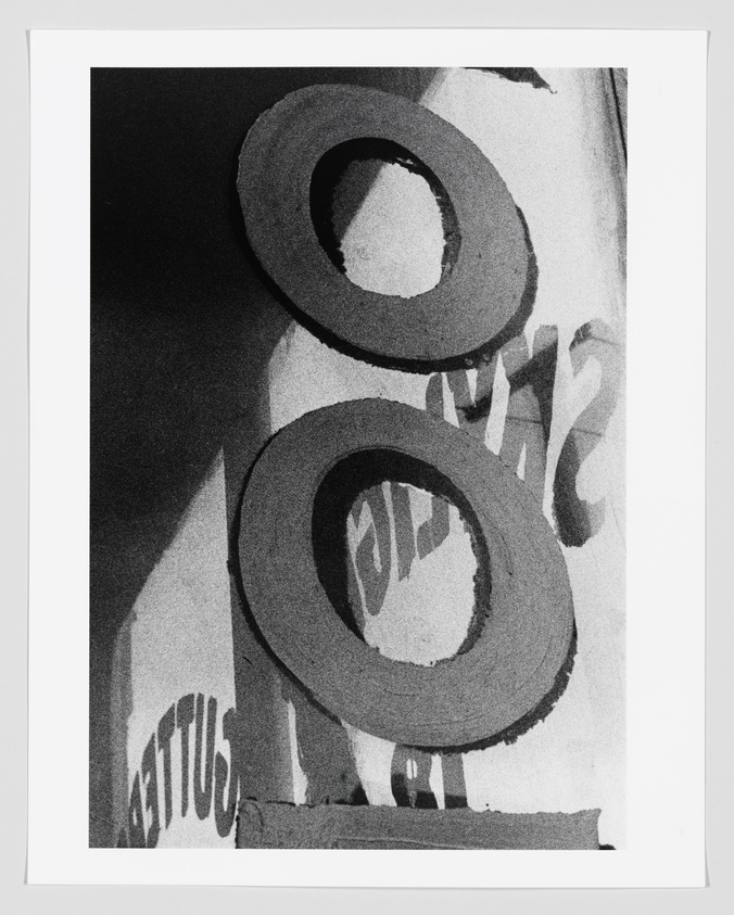 A black and white abstract photo featuring large, bold numbers or letters with a partial view of a person's face in the background. The text appears to be reversed, with "GUTTUS" partially visible, suggesting the image may be a reflection or taken through transparent material with writing on it.