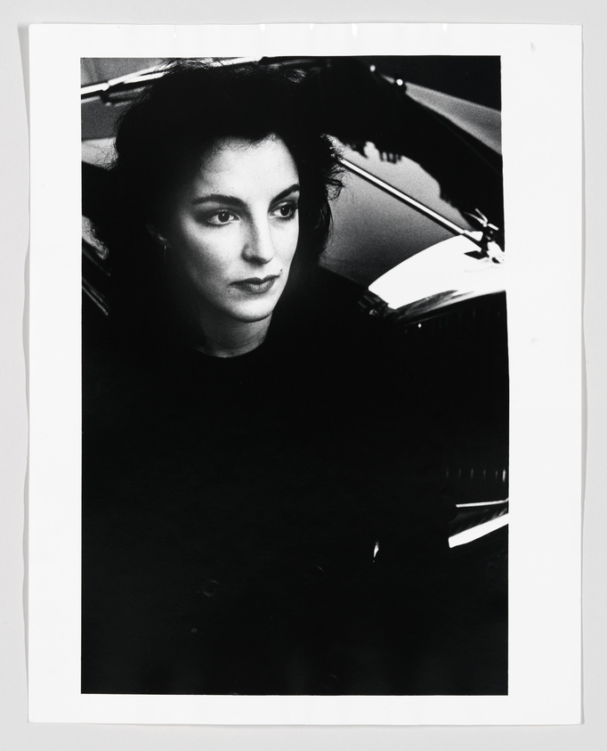 A black and white photograph of a woman with dark hair looking off to the side, seated in a car with the interior and part of the hood visible in the frame. The image has high contrast with deep shadows and bright highlights.