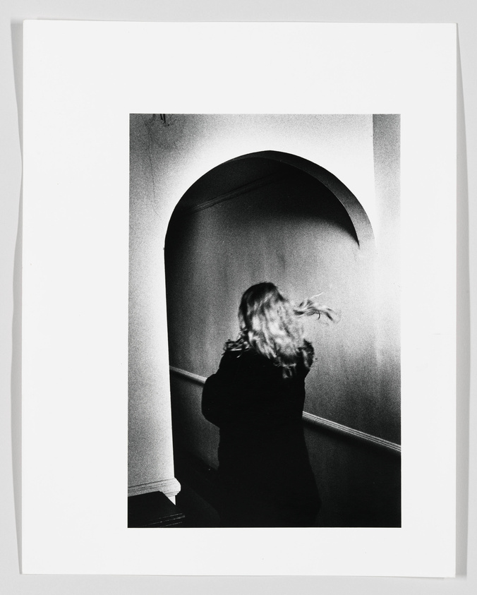 A black and white photograph capturing a person with long hair from behind as they walk through an arched passageway. The person's hair is caught in motion, giving a sense of movement within the still image. The contrast between light and shadow adds depth to the scene.