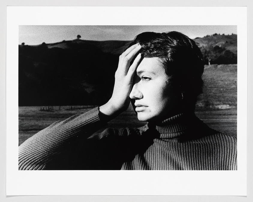 A black and white photograph of a person in profile with their hand on their forehead, wearing a ribbed turtleneck sweater. The background features a hilly landscape under a clear sky.