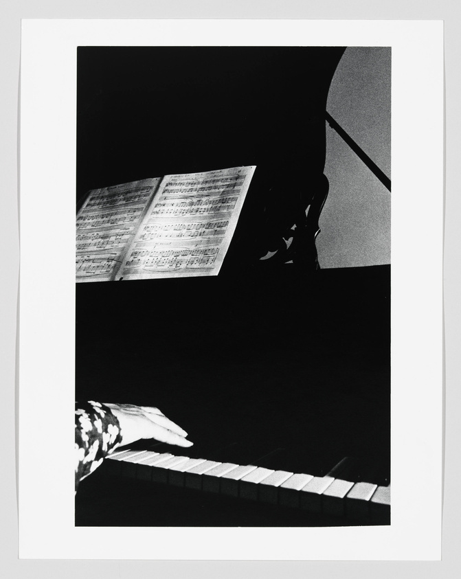 A high-contrast black and white photograph capturing a musician's hand playing a piano with sheet music visible in the background. The image focuses on the hand, piano keys, and the sheet music, with the rest of the scene shrouded in darkness.