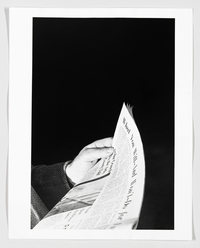 A black and white photograph of a person's hand holding an open newspaper, with the text on the paper partially visible and the rest of the image surrounded by a white border.