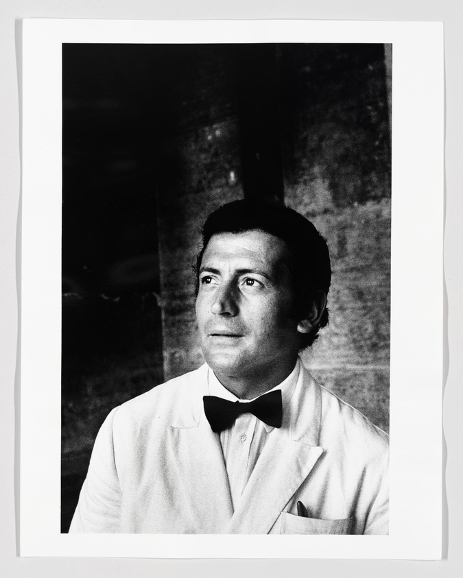 Black and white portrait of a man in a white jacket and bow tie looking off to the side with a thoughtful expression. The background is dark and indistinct, highlighting the subject's face.