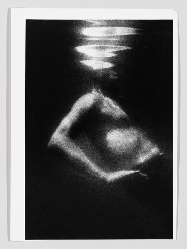 A black and white photograph depicting a blurred figure of a person submerged in water, with light patterns reflecting on the surface above them. The image has a dreamlike, ethereal quality due to the motion blur and high contrast.