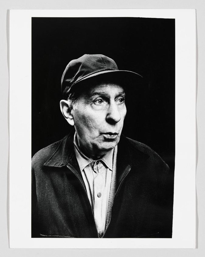 Black and white portrait of an elderly man wearing a cap and a jacket over a buttoned shirt, looking to the side with a thoughtful expression.