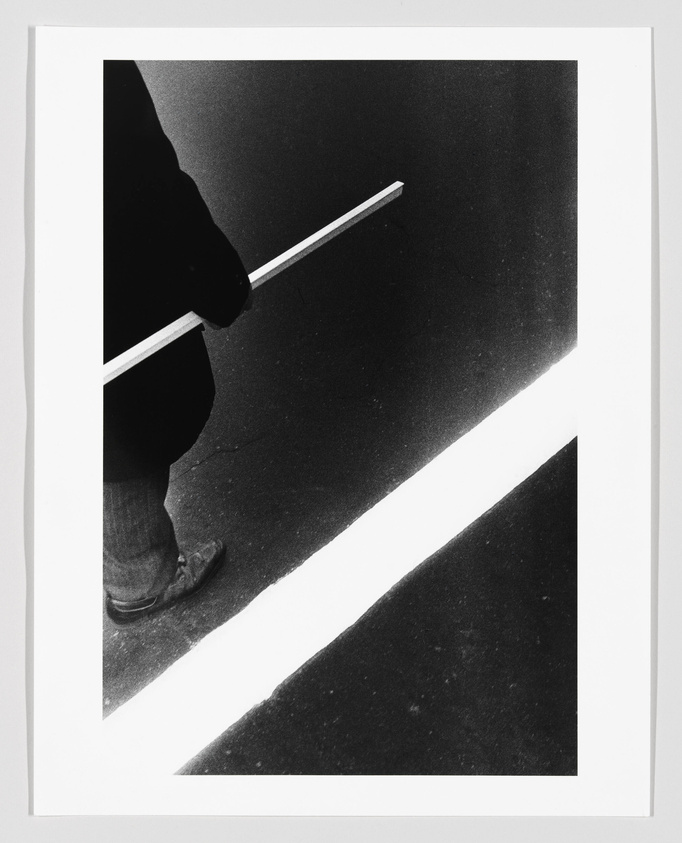 A high-contrast black and white photograph capturing a partial view of a person's lower body standing next to a bright white line on the ground, with a cane or stick extending over the line. The image is framed to emphasize the stark contrast between light and shadow.