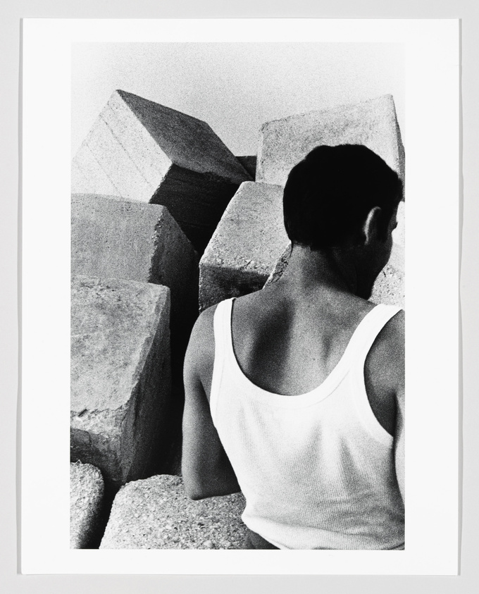 A black and white photograph of a person from behind, wearing a white tank top, looking at large geometric concrete blocks.