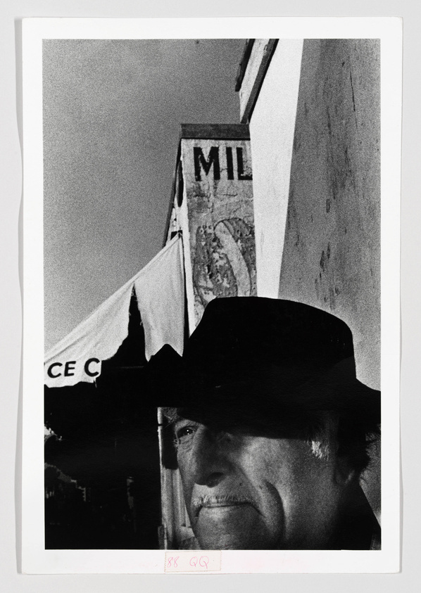 A black and white photograph featuring a close-up of a person wearing a wide-brimmed hat, with a partial view of a building and a flag with the letters "CE C" visible in the background. The image has a grainy texture, and there are handwritten markings at the bottom.