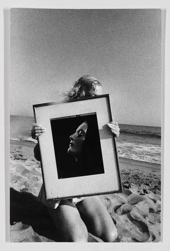 A black and white photograph of a person sitting on a sandy beach holding a framed portrait in front of their face, aligning the portrait's face with their own body. The ocean and waves are visible in the background, and the image has a grainy texture.