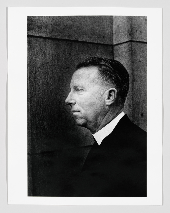 Black and white photograph of a man in profile with a clean-cut appearance, wearing a dark suit and a white shirt. The background features a textured wall, and the image is framed with a white border.