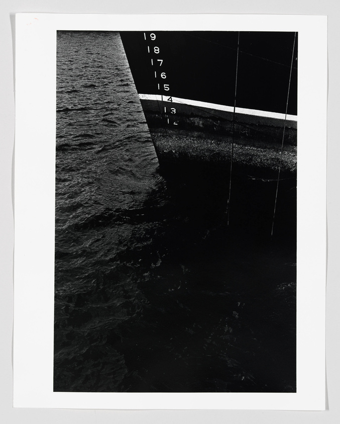A black and white photograph showing a close-up of a ship's hull with draft marks numbered from 12 to 19, partially submerged in water, with the sea's surface visible on the left side of the image.