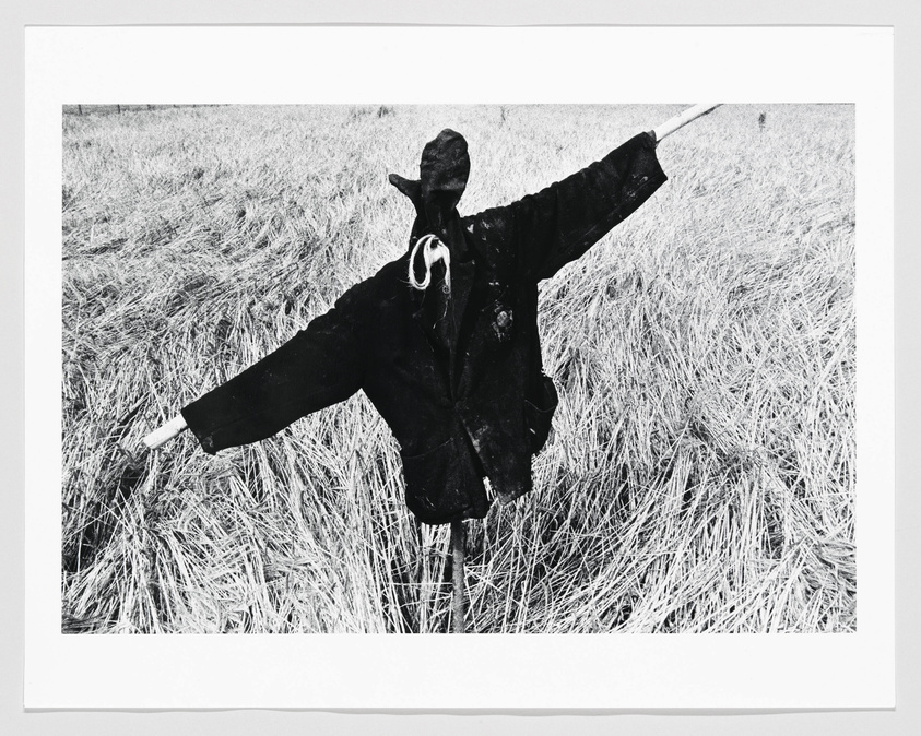 A black and white photograph of a scarecrow wearing a dark coat with outstretched arms in a field of dry grass.