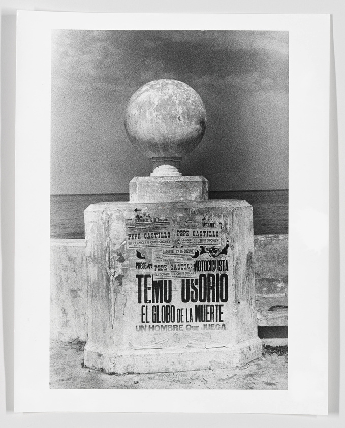 A black and white photograph of a weathered monument with a large spherical object on top, against a cloudy sky and calm sea backdrop. The monument has various posters with text, including the prominent words "TEMERARIO EL GLOBO DE LA MUERTE" and "PEPE CASTILLO".